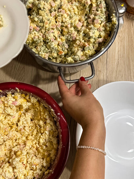 Close-up of a hand preparing traditional Russian salad (Olivier salad) in a home setting. Large bowls of mixed ingredients, including potatoes, eggs, and ham, are displayed on a wooden table.