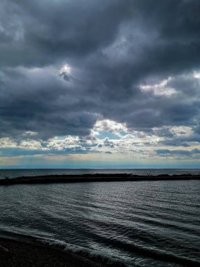 Dark, dramatic storm clouds loom over a calm ocean with subtle light breaking through. The photo captures a moody seascape ideal for themes of nature, weather, and introspection. clipart