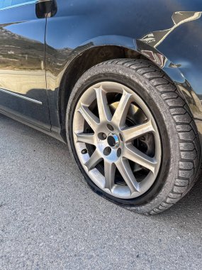Şehir caddesinde park edilmiş siyah bir arabanın ön lastiği patlamış. Sönük tekerleğe yakın çekim. Araba arızalarını, araba tamirlerini, trafik kazalarını ve şehir içi araç sorunlarını resmetmeye elverişli.