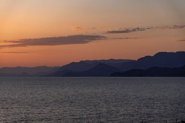 Kıyı dağları, renkli bir günbatımı gökyüzünün altında akşam sisi içinde kaybolur. Tasarım ve web sitelerinde seyahat, farkındalık, meditasyon veya arka plan kullanımı için ideal sakin deniz manzarası.