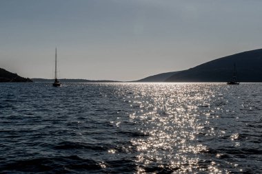 Yelkenliler, parlak su yansımaları ve alacakaranlıktaki uzak tepelerle güneşli bir denizde sürüklenirler. Romantik, denizcilik, özgürlük, seyahat ve ilham verici temalar için ideal.