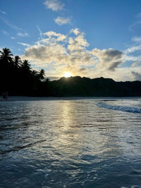 Sahilde baş döndürücü bir günbatımının sakin görüntüsü, suyun üzerinde altın yansımalar ve gökyüzüne yayılan yumuşak bulutlar. Anahtar sözcükler: sükunet, doğa ve manzaranın güzelliği.