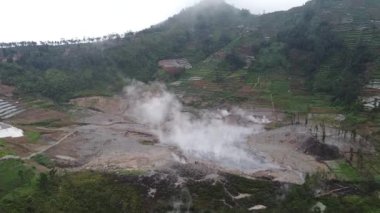 Yerden yükselen buharlı, teraslı tarlalar ve tepelerle çevrili jeotermal bir alanın havadan görüntüsü. Manzara, arka planda çorak alanlar ve bitki örtüsüyle volkanik aktivite belirtilerini gösteriyor..