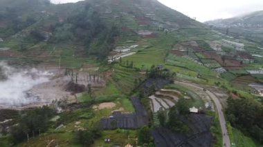 Yamaçtaki tarımsal tarlaların havadan görünüşü, yeşil ekinler ve koyu plastik örtüler. Manzara virajlı yollar ve sisli bir atmosfere sahiptir. Dieng Platosu.