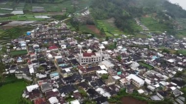 Teraslı tarlalar ve tepelerle çevrili yoğun nüfuslu bir köyün hava manzarası. Köy, merkezinde tanınmış bir cami bulunan geleneksel ve modern mimarinin bir karışımına sahip. Manzara yemyeşil.