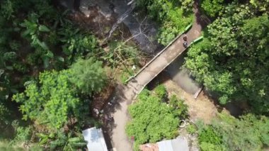 Derenin üzerinde küçük bir köprü bulunan yemyeşil bir arazinin havadan görünüşü. Bölge ağaçlar ve çalılar da dahil olmak üzere yoğun bitki örtüsüyle çevrili ve köprüye giden toprak bir yol var..