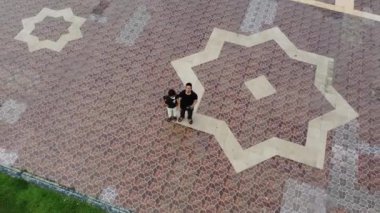 Geometrik tasarımları olan havadan bir kaldırım manzarası. Otlak bir alanda köpeği gezdiren bir kişi. Karmaşık fayans desenleri yeşil çimlerle görsel olarak çekici bir kontrast oluşturuyor..