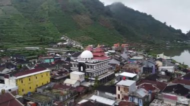 Arka planda tarlaları olan bir köyün havadan görünüşü. Ön planda, merkezinde tanınmış bir cami bulunan geleneksel ve modern evlerin bir karışımı yer alıyor. Dieng Platosu, Endonezya.