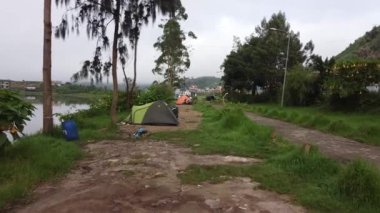 Göl kenarındaki kamp alanının manzarası. Otlak bir yola kurulmuş birkaç çadır var. Manzara, arka planda sisli bir atmosfere sahip ağaçlar ve bir patika içerir. Dieng Platosu, Endonezya.