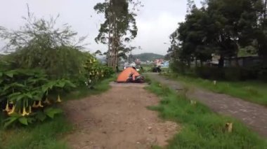 Göl kenarındaki kamp alanının manzarası. Parlak turuncu bir çadır ve park etmiş bir scooter. Yeşillik, yolu çevreleyen ağaçlarla çevrili. Dieng Platosu, Endonezya