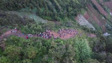 Teraslı, yemyeşil bir arazinin havadan görünüşü. Büyük bir grup insan yoğun bitki örtüsüyle çevrili bir toprak yolda toplanıyor. Sahne Dieng 'in güzelliğini gözler önüne seriyor..