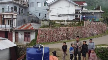 Wonosobo, Endonezya - 12132021: Bir grup çocuk ve yetişkin, evleri ve su tanklarıyla çevrili kırsal bir alanda toplanıyorlar. Sahne, çocukların gülümseyip oynadığı neşeli bir anı yakalıyor.