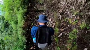 Mavi şapkalı ve sırt çantalı bir adam yemyeşil yollarla çevrili dar bir patikada yürüyüş yapıyor..