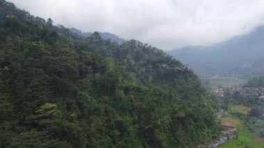 Yoğun ormanlar ve tepelerin üzerinde süzülen sisli bulutlarla dolu yemyeşil dağlık bir manzara. Sahne, aşağıdaki vadide görünen küçük bir köyle tepelerin doğal güzelliğini yakalar..