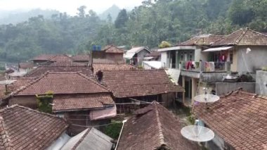 Kiremit çatılı geleneksel evleri olan kırsal bir köyün havadan görünüşü. Manzara yemyeşil, tepeler ve dağlarla çevrili. Bazı evlerin çamaşırları dışarıda asılı ve uydu antenleri çatılarda görülebiliyor..