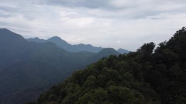 Bulutlu bir gökyüzünün altında yemyeşil ormanlarla dolu bir dağ manzarası..