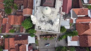 Etrafı ağaçlar ve binalarla çevrili iki minareli bir caminin havadan görünüşü. Cami, birkaç kişinin oturduğu düz bir çatı ve avlu ve yakınlarda park edilmiş bir araba barındırıyor..
