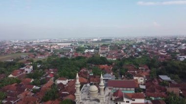Kırmızı çatılı evleri ve yeşil bitkileri olan yoğun nüfuslu bir yerleşim bölgesinin havadan görünüşü. Ufuk, temiz bir gökyüzü altında kentsel ve kırsal manzaraların bir karışımını gösteriyor..