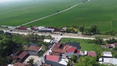 Evleri, yolları ve geniş yeşil arazileri olan kırsal bir bölgenin havadan görünüşü. Manzara, yola paralel uzanan küçük bir gölet ve bir kanal içerir..