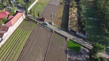 Küçük bir yolu ve kırmızı bir aracı olan tarımsal tarlaların havadan görünüşü. Tarlalar çeşitli tarım safhalarında, bazı bölgeler yeni sürülmüş, diğerleri de yeşil mahsulle sergileniyor..