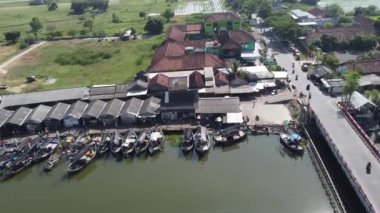 Nehir boyunca kenetlenmiş çok sayıda teknesi olan hareketli bir balıkçı köyünün havadan görüntüsü. Köyde geleneksel ve modern binaların karışımı var, arka planda yeşil alanlar ve su kaynakları var..