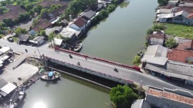 Her iki tarafında evler olan bir yerleşim bölgesindeki nehrin üzerindeki küçük bir köprünün havadan görüntüsü. Motosikletler köprüden geçiyor ve tekneler nehir kıyısına yanaşıyor..