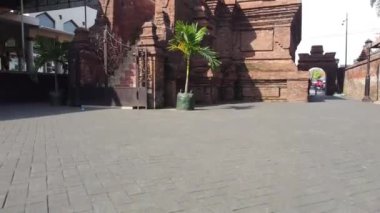 Karmaşık mimari ayrıntılara sahip tarihi bir tuğla bina, asfaltlı bir caddede yer alan kapılı bir giriş ve saksı bitkileri yer alıyor. Menara Kudus.