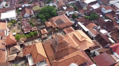 Kırmızı kiremitli çatıları olan yoğun bir yerleşim bölgesinin havadan görüntüsü. Evler yakın inşa edilmiş ve geleneksel ve modern yapıların bir karışımı var. Çevreye yeşil ağaçlar saçılmış. Menara Kudus.