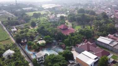 Yüzme havuzu ve yeşilliği olan yerleşim alanının havadan görünüşü. Görüntü kırmızı çatılı evleri, ağaçları ve yüzen insanlarla dolu bir havuzu gösteriyor. Arka planda daha çok ev ve puslu bir gökyüzü var..