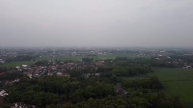 Yoğun yeşillik, ağaçlar ve bulutlu bir gökyüzünün altında dağınık evler olan kırsal bir arazinin havadan görünüşü..