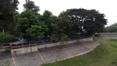 Önünde ağaçlarla ve arka planda bir sıra küçük yapı veya tezgahlarla sıralanmış çimenli bir alan olan beton bir set..