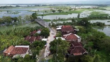 Kırmızı çatılı evleri olan, yemyeşil tarlalar ve su kaynaklarıyla çevrili kırsal bir köyün havadan görünüşü. Köyün içinden geçen bir yol, nehrin üzerindeki bir köprüye çıkıyor..