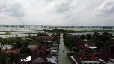 Sular altında evler ve tarlalarla dolu bir köyün havadan görünüşü. İnsanlar teknelerle sel sularında yüzüyorlar. Gökyüzü bulutlu ve manzara çoğunlukla ağaçlar ve elektrik hatlarıyla düz..
