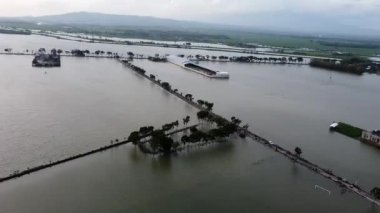 Su altında kalmış yollar, evler ve tarlaların bulunduğu su basmış bir bölgenin havadan görüntüsü. İnsanlar kısmen sular altında kalmış bir yolda yürürken görülüyor. Gökyüzü kapalı..
