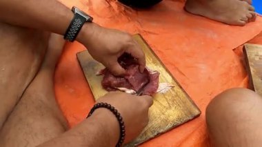 Tahta kesme tahtasıyla çiğ et doğrayan birinin yakın çekimi. Kişi bilezik ve saat takıyor. Arka plan turuncu bir yüzey ve arka planda başka bir kişi görünüyor..