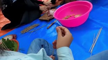 İnsanlar mavi bir hasırda yemek hazırlıyorlar, içinde şişler ve pembe bir kase var..