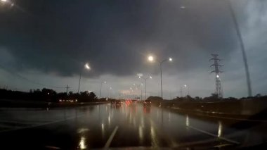 Gece ıslak bir yolda giden arabaların ışıkları manzarayı aydınlatıyor ve tepemizde bulutlu, karanlık bir gökyüzü var..