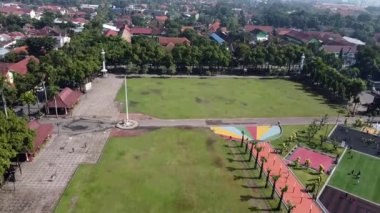 Kentsel bir alanda ağaçlar ve binalarla çevrili geniş bir açık arazinin havadan görüntüsü. Alanın ortasında bir bayrak direği ve bir tarafında renkli bir bölüm var. Jombang, Doğu Java, Endonezya.