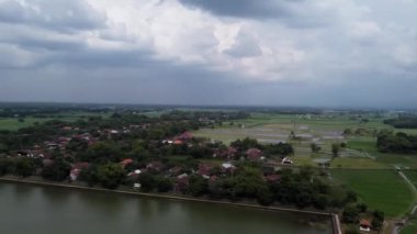 Endonezya, Mojokerto 'da Majapahit kraliyet havuzu. Yeşillik ve tarlalarla çevrili, bulutlu bir gökyüzünün altında bir su kütlesine bitişik, kırsal bir köyün havadan görünüşü..