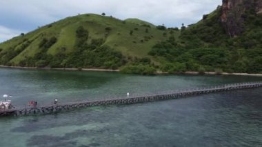 Manjarite Sahili, Labuan Bajo: Berrak ve sakin suları yemyeşil yamaçlara doğru uzanan uzun bir ahşap iskele manzarası. İnsanlar rıhtımda yürüyor, doğal çevrenin tadını çıkarıyorlar.