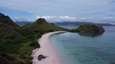 Komodo Adası 'ndaki Pembe Plaj, Labuan Bajo, Endonezya: Arkaplanda dağlık bir ada bulunan sakin ve berrak bir mavi denizde yüzen iki teknenin havadan görünüşü parçalı bulutlu bir gökyüzünün altında.