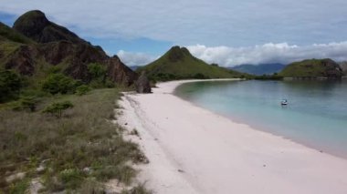 Komodo Adası 'ndaki Pembe Plaj, Labuan Bajo, Endonezya: Pembe kumlu sakin bir plaj, berrak turkuaz su, ve arka planda yemyeşil tepeler. Kıyıya yakın küçük bir tekne demir atmış..