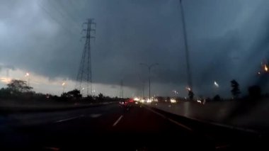 Yoğun bir otoyolun üzerinde karanlık, fırtınalı bir gökyüzü. İki yönde de giden arabalar var. Sokak lambaları yanıyor ve yaklaşan fırtınayla atmosfer gergin görünüyor..