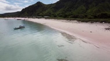 Labuan Bajo, Endonezya: Temiz turkuaz suyu olan sakin bir sahil, kıyıya demirli küçük bir tekne ve arka planda yemyeşil tepeler. Sahilde birkaç kişi görülüyor..