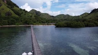 Labuan Bajo, Endonezya - 03 08 2023: Berrak mavi suların üzerinden yemyeşil tepelere doğru uzanan ahşap bir köprünün manzarası. Beyaz şemsiyeli bir tekne köprüye yanaştı..