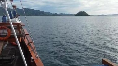 Labuan Bajo 'daki bir teknenin manzarası sakin suları ve dağlarla çevrili uzak adaları gösteriyor. Gökyüzü parçalı bulutlu, sakin bir atmosfer yaratıyor..