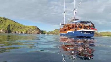 Labuan Bajo 'nun sakin sularına demir atmış, yemyeşil tepeler ve kayalık kayalıklarla çevrili ahşap bir yelkenli gemi. Gökyüzü parçalı bulutlu, sakin bir atmosferi yansıtıyor..