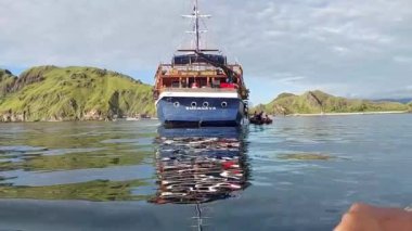 Labuan Bajo 'daki yemyeşil bir adanın yakınında demirleyen büyük bir tekne sakin sulara yansıyor. Gökyüzü parçalı bulutludur ve manzara arka planda kayalık tepeler barındırır..