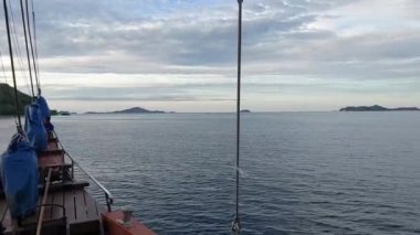 Labuan Bajo 'daki bir teknenin güvertesinden manzara uzak adalara doğru sakin sulara bakıyor. Gökyüzü yumuşak ışıkla parçalı bulutlu, sakin bir atmosfer yaratıyor..