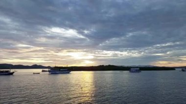 Labuan Bajo 'da sakin bir denizin üzerinde sakin bir günbatımı, ön planda demirli tekneler ve uzakta adalar. Gökyüzü sıcak turuncu renkleri yansıtan bulutlarla dolu..